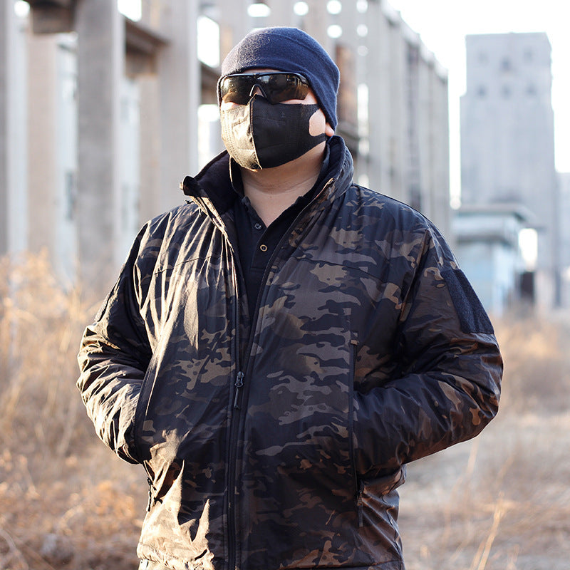 A person wearing a camouflage waterproof coat with a hood, sunglasses, and a beanie. They are standing in a dilapidated building setting.