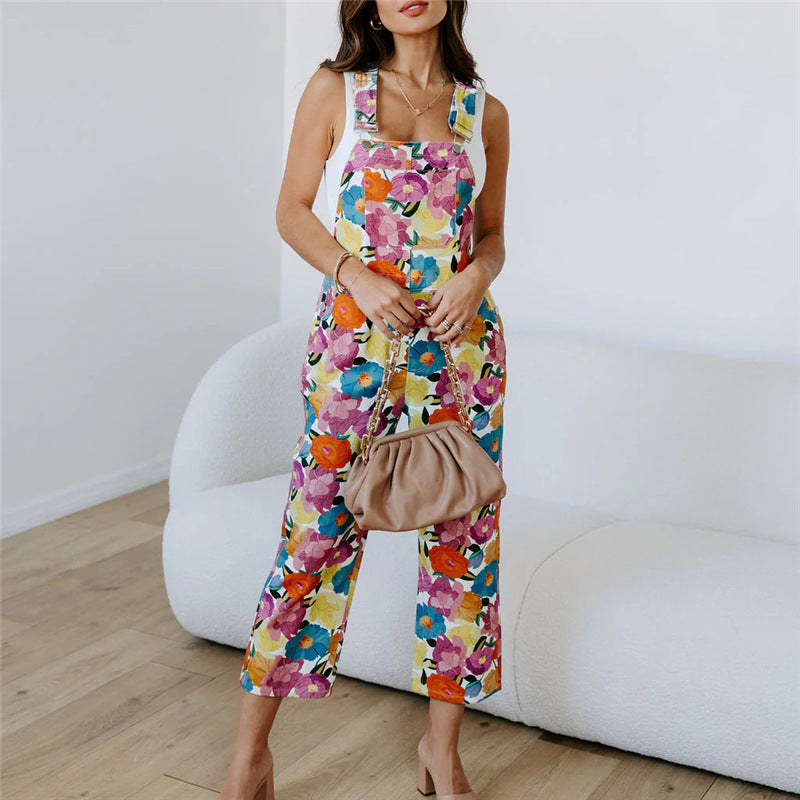 A model wearing a sleeveless, straight-leg floral print jumpsuit in a room with a white background.