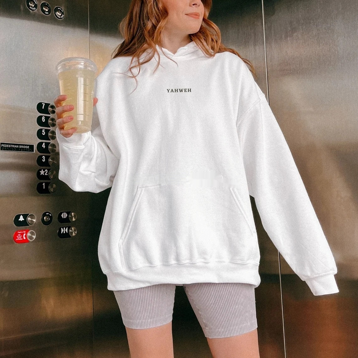 A woman wearing a white hoodie with a bold black 3D letter print, paired with gray bicycle shorts, standing inside an elevator.