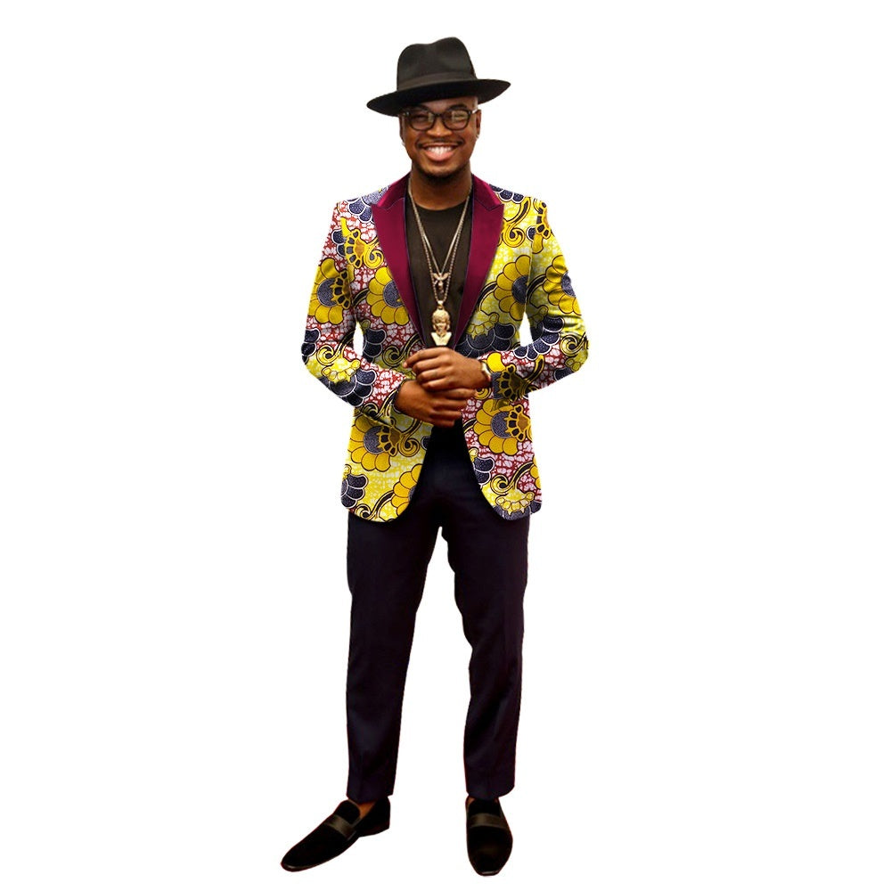 A man wearing a multicolored patterned blazer with a peak lapel, long sleeves, and a slim-fit design. He is also wearing a hat and glasses and is posed against a plain background.