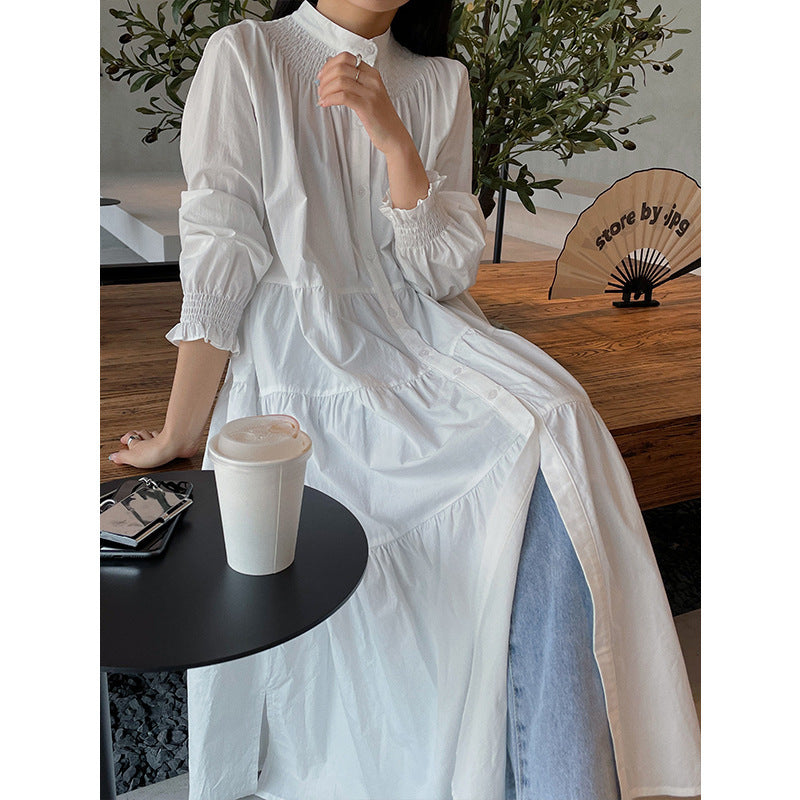 A woman sitting at a table wearing a white midi-length pleated dress with a round neckline and long sleeves.