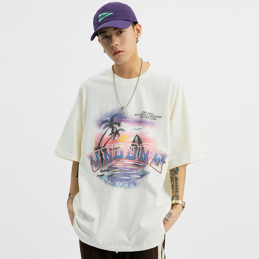A person wearing a white short-sleeved t-shirt with a seaside landscape print, including a palm tree and a sunset, paired with dark pants and a purple cap.