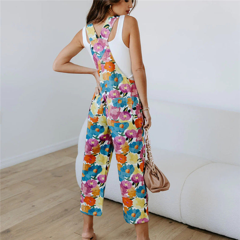 A model wearing a sleeveless, straight-leg floral print jumpsuit in a room with a white background.