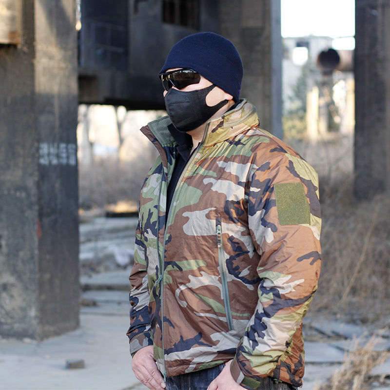 A person wearing a camouflage waterproof coat with a hood, sunglasses, and a beanie. They are standing in a dilapidated building setting.