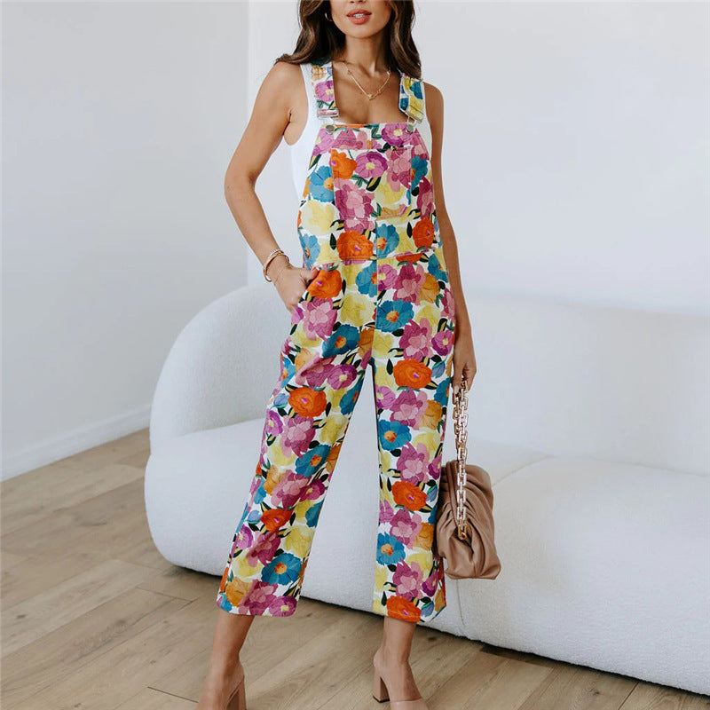A model wearing a sleeveless, straight-leg floral print jumpsuit in a room with a white background.