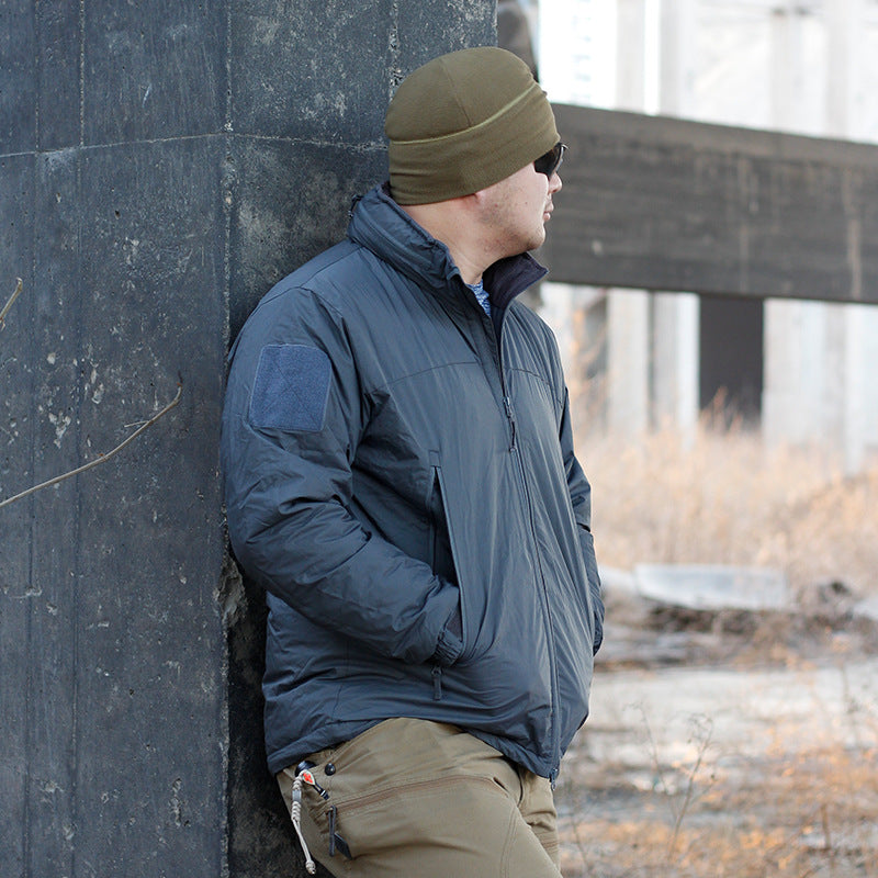 A person wearing a camouflage waterproof coat with a hood, sunglasses, and a beanie. They are standing in a dilapidated building setting.