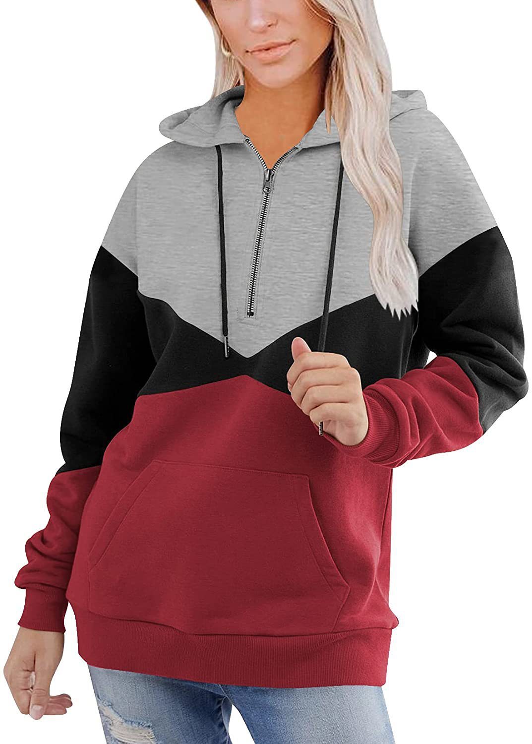 A woman wearing a color-blocked hoodie with a hood, in beige, black, and green colors, holding a phone and smiling.