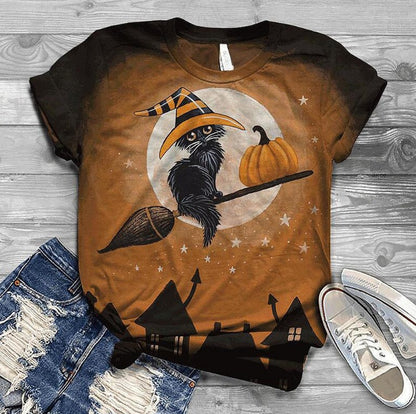 Halloween-themed t-shirt with a crow, pumpkin, and broomstick design on a wooden surface with jeans and shoes.